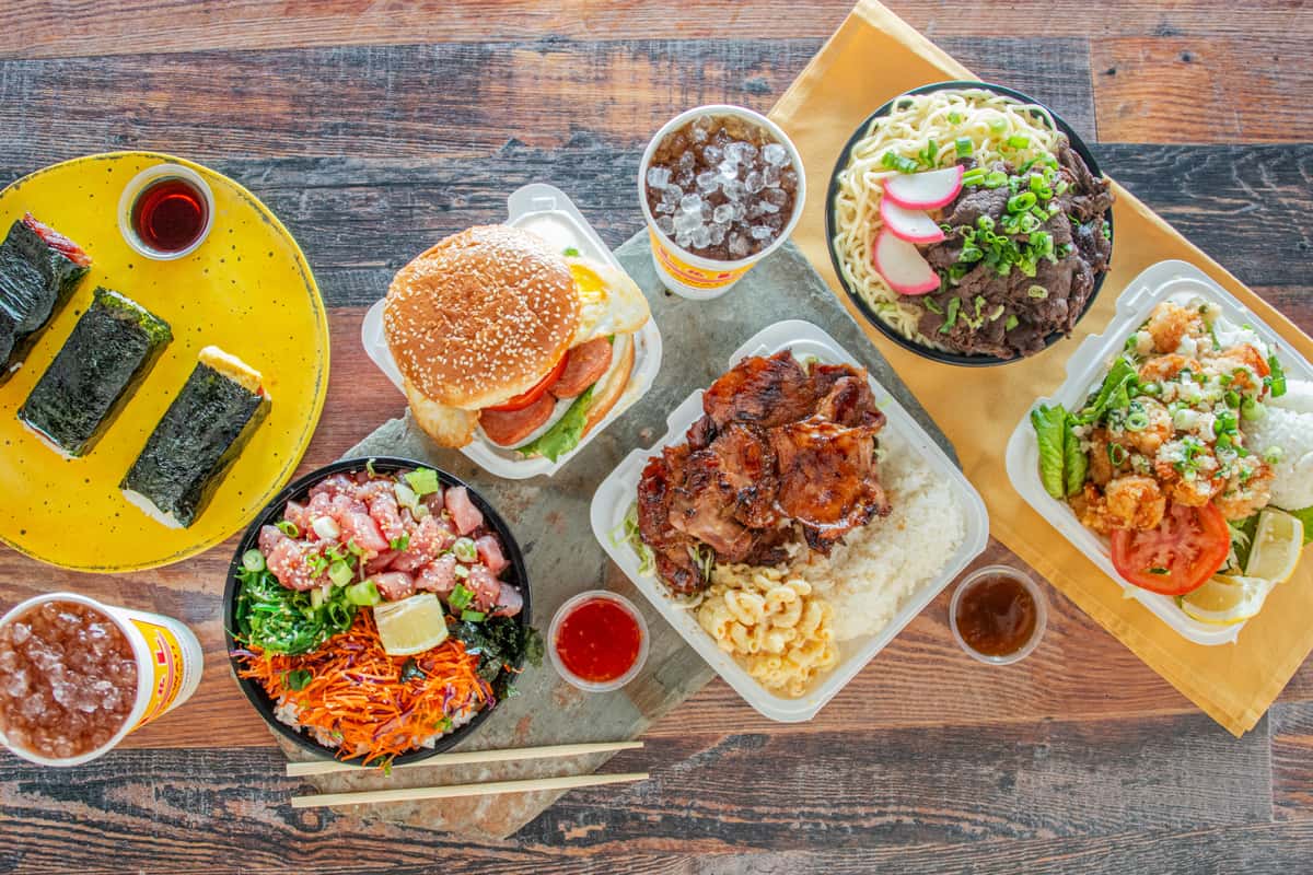 Overhead view of Hawaiian plate lunch spread with poke bowl, BBQ chicken, musubi, beef ramen, and shrimp plate on wood table