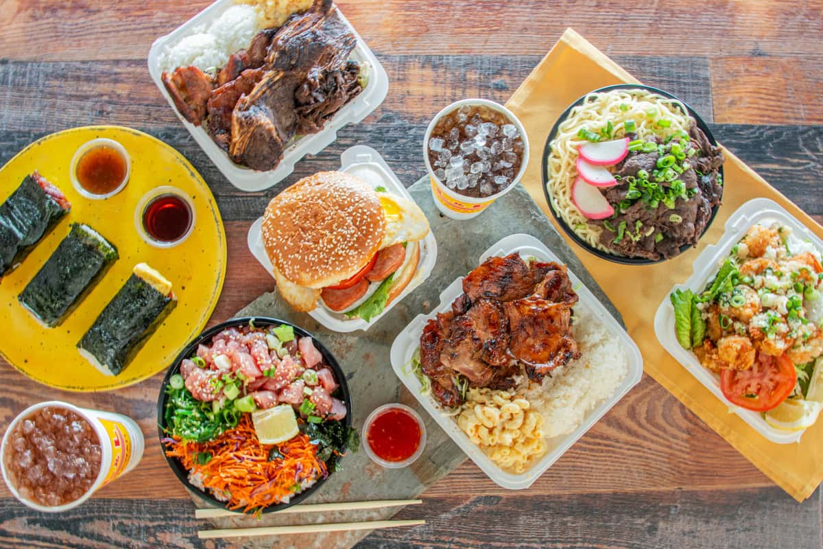 Overhead view of Hawaiian plate lunch spread with poke bowl, BBQ chicken, musubi, beef ramen, and shrimp plate on wood table
