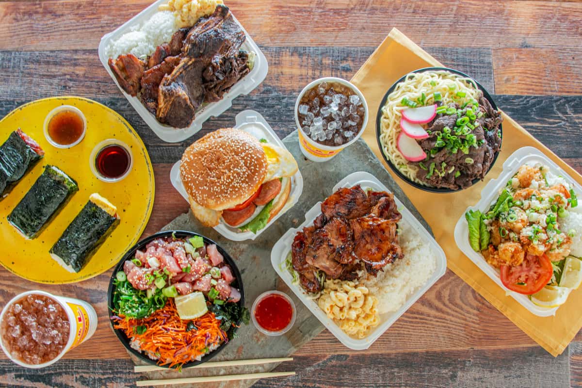 Overhead view of Hawaiian plate lunch spread with poke bowl, BBQ chicken, musubi, beef ramen, and shrimp plate on wood table