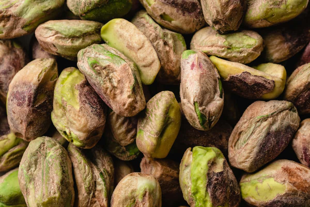 This image shows a close-up of shelled pistachio nuts. The nuts display a natural mix of green and purplish-brown hues, highlighting their raw, unprocessed state. The texture appears dry and slightly wrinkled, typical of pistachios with the skin still on.