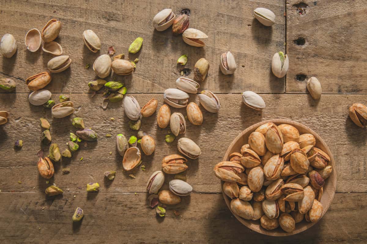 Scattered pistachios and cracked shells on a rustic wooden table with a wooden bowl full of roasted pistachios on the right.