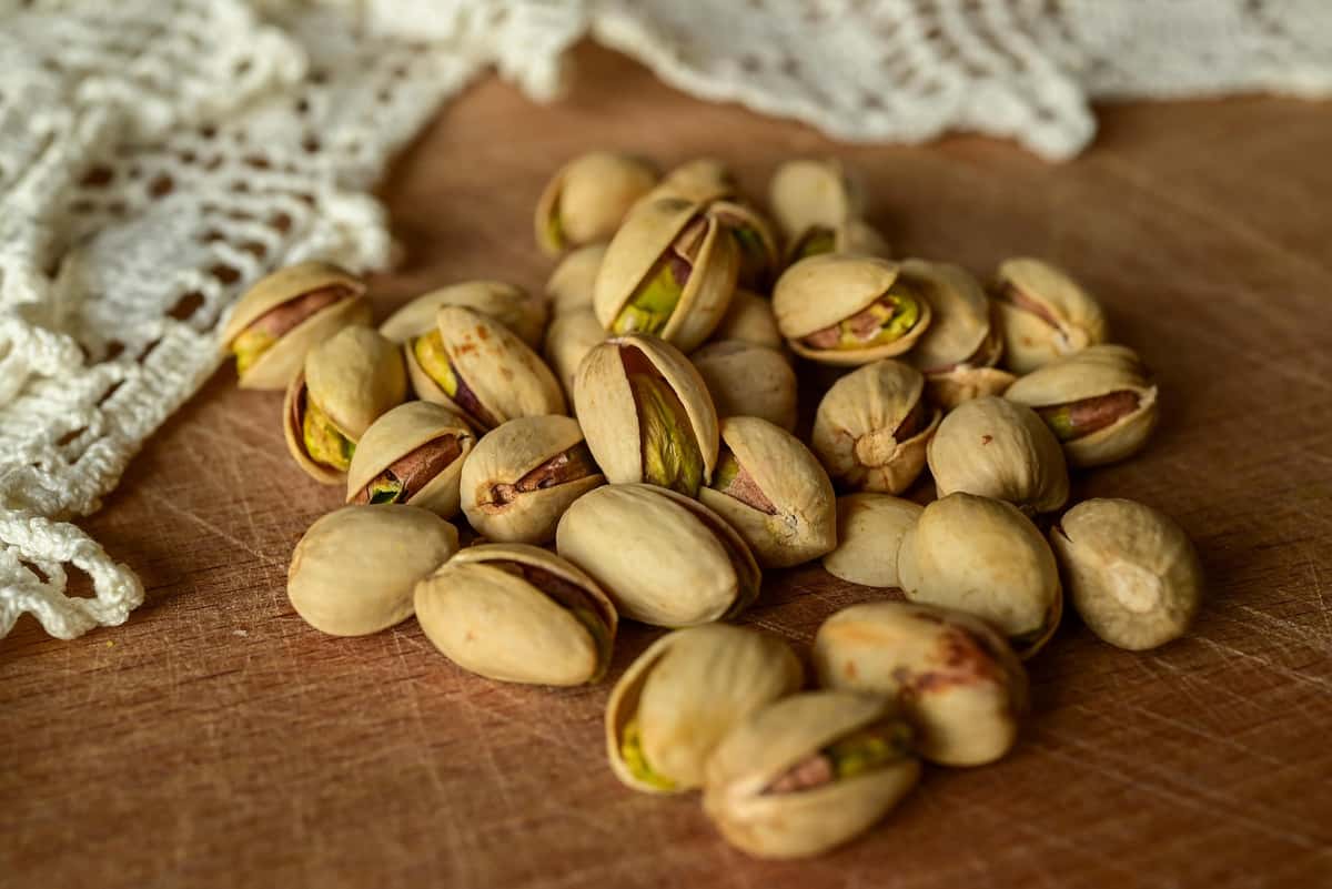 A pile of roasted pistachios in shells on a wooden surface next to a white crocheted cloth.