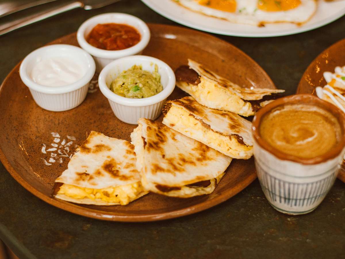 Grilled breakfast quesadillas with scrambled eggs, served with guacamole, salsa, sour cream, and a frothy coffee on the side.