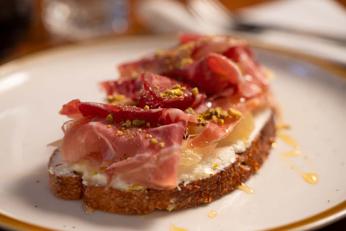 A slice of toasted bread topped with cream cheese, prosciutto, pistachios, and a drizzle of honey.