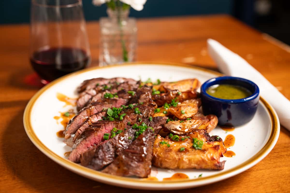 A plate of medium-rare sliced steak served with roasted potatoes, green onions, and a small bowl of dipping sauce. A glass of red wine accompanies the dish.