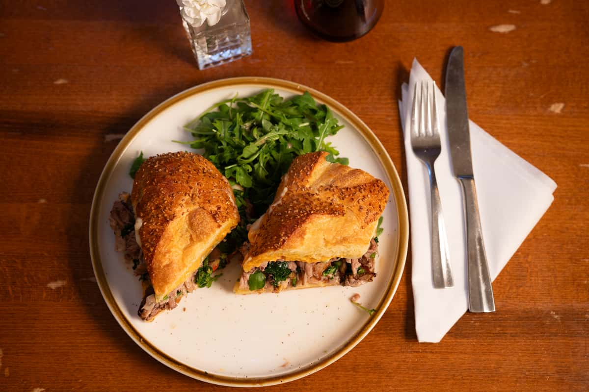 A sandwich with sliced beef, arugula, and herbs in a sesame seed bun, served with a side of arugula salad.
