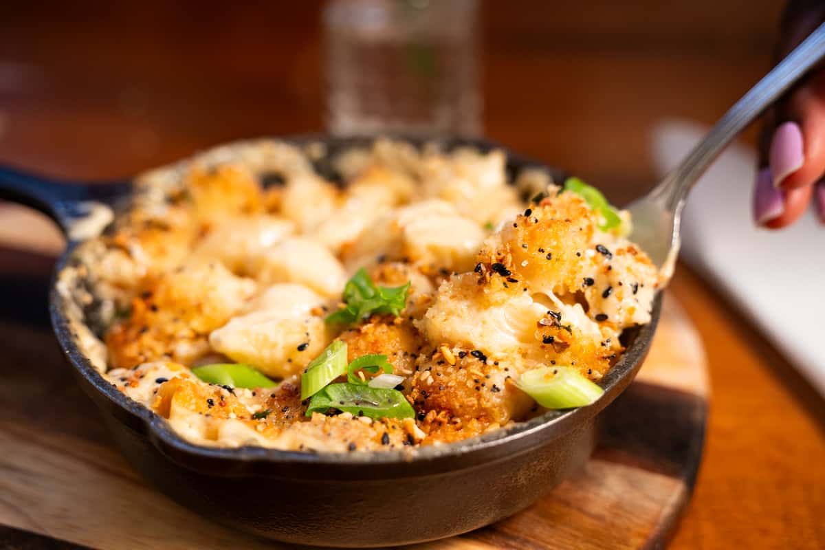 A spoon lifts a serving of creamy baked macaroni and cheese with a crunchy topping, garnished with green onions, from a cast-iron skillet.