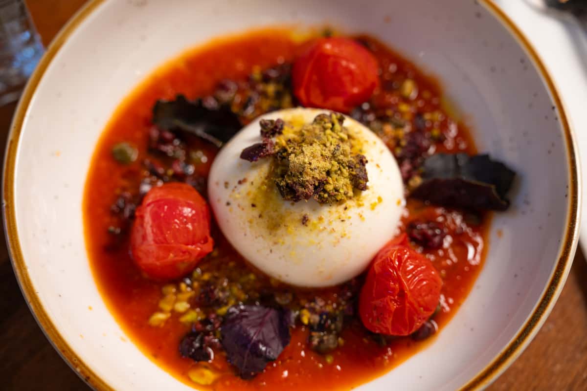 A serving of burrata cheese sits in a tomato sauce with roasted cherry tomatoes, garnished with pistachios and olives.