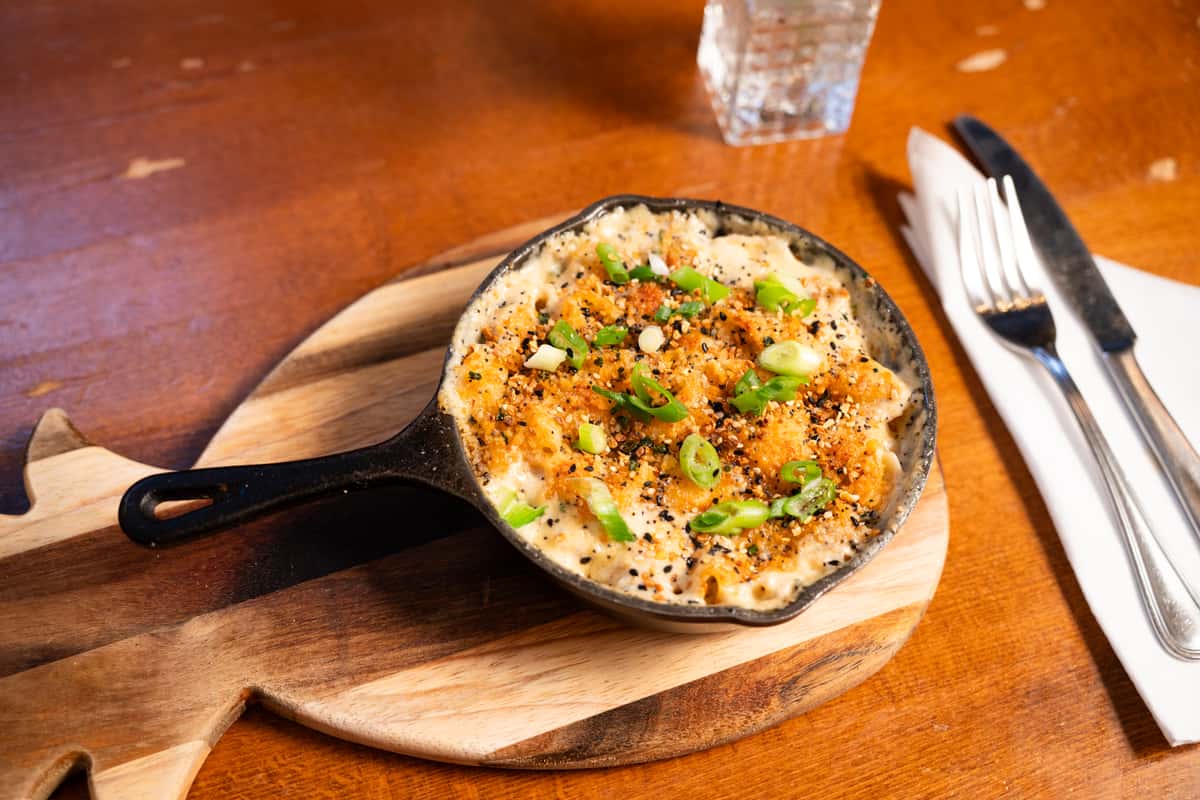 A cast iron skillet filled with baked macaroni and cheese, topped with breadcrumbs and green onions, served on a wooden board. A fork and knife are beside the dish.