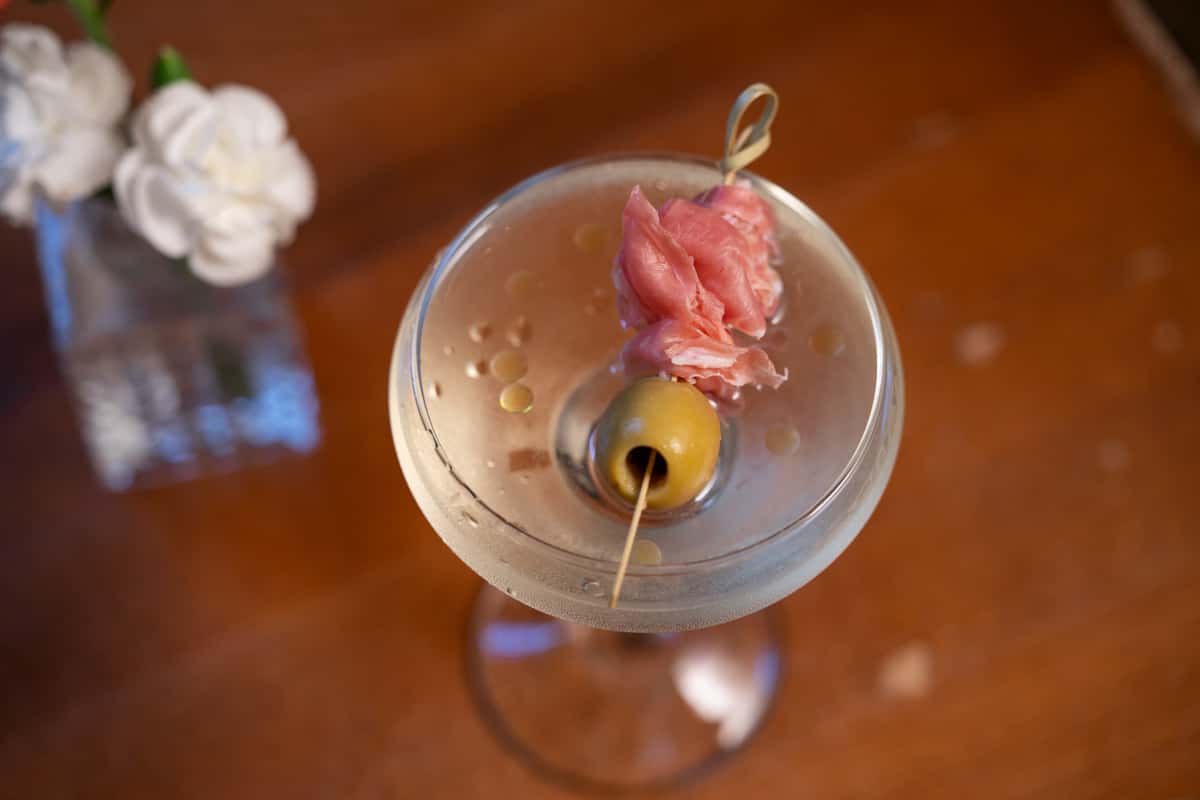 A classic martini garnished with an olive and a slice of prosciutto on a cocktail pick, served in a chilled glass.