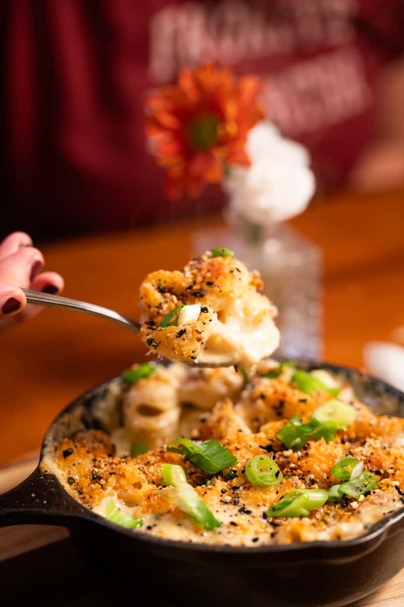 A spoon lifts a serving of creamy baked macaroni and cheese with a crunchy topping, garnished with green onions, from a cast-iron skillet.