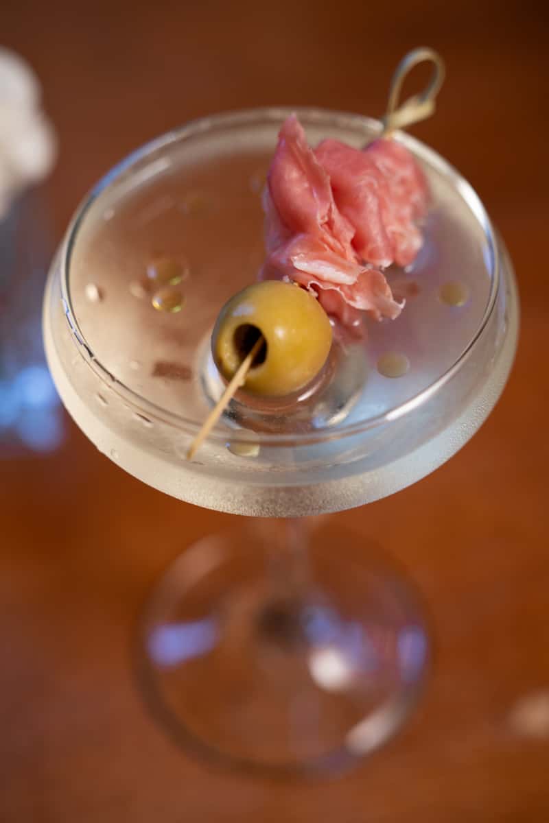 A classic martini garnished with an olive and a slice of prosciutto on a cocktail pick, served in a chilled glass.