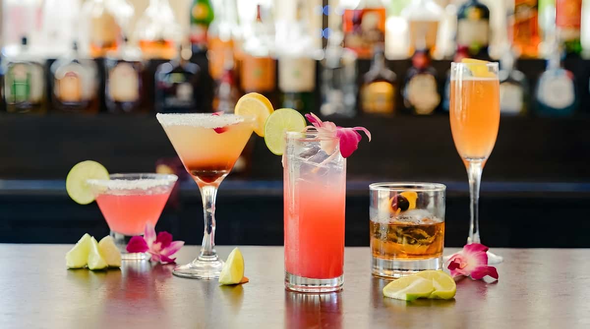 Five colorful cocktails and lime/lemon wedges on a smoky bar counter with blurred liquor bottles in background.