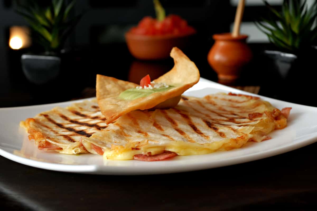 Grilled ham and cheese quesadilla with melted cheese, topped with a crispy tortilla chip and creamy avocado sauce, served on a white plate in a cozy restaurant setting.
