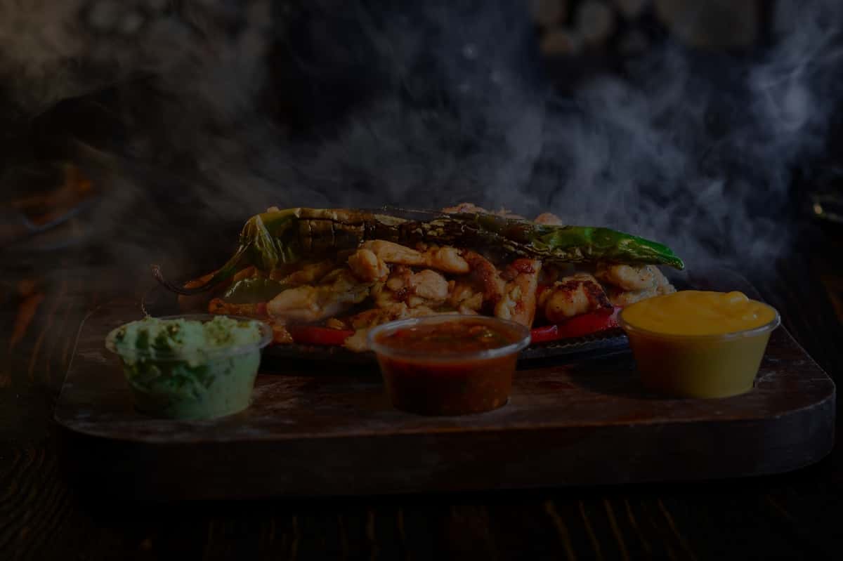 Delicious sizzling fajitas served with guacamole, salsa, and cheesy sauce on a wooden tray.