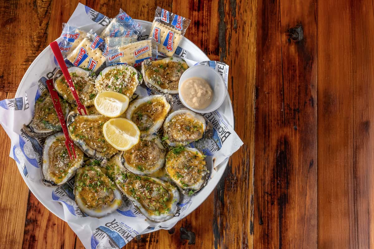 Chargrilled oysters topped with garlic butter, parmesan, and herbs, served with lemon wedges, saltine crackers, horseradish, and red cocktail forks on branded Backwater Grill paper.