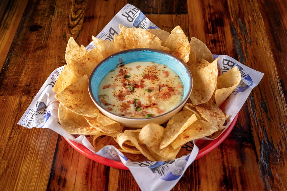 Warm queso dip topped with diced tomatoes, jalapeños, and seasoning, surrounded by crispy tortilla chips dusted with chili powder, served in a red basket lined with Backwater Grill paper.