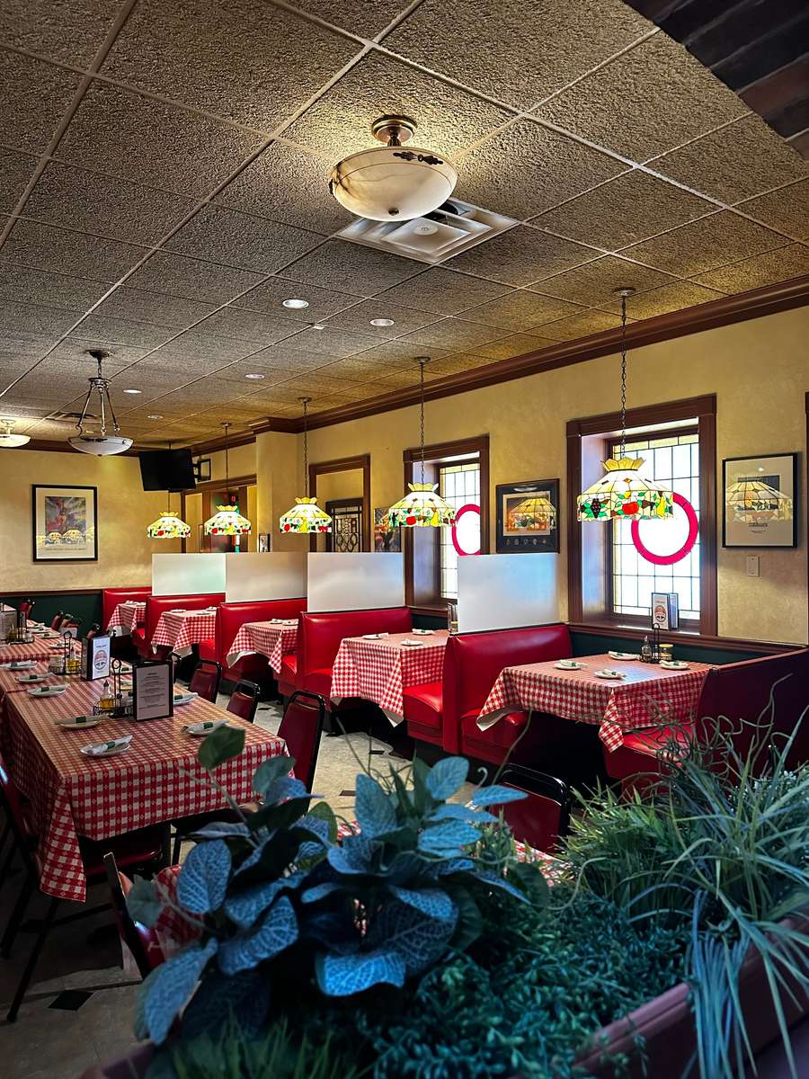 Cozy Italian restaurant interior featuring red leather booths, checkered red-and-white tablecloths, stained glass pendant lights, and framed vintage artwork, creating a warm and nostalgic dining ambiance.