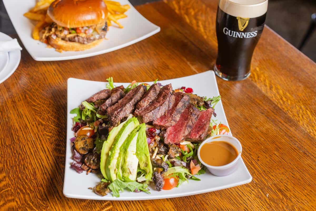 Sliced grilled steak salad with fresh avocado, mixed greens, and house dressing served on a wooden table.