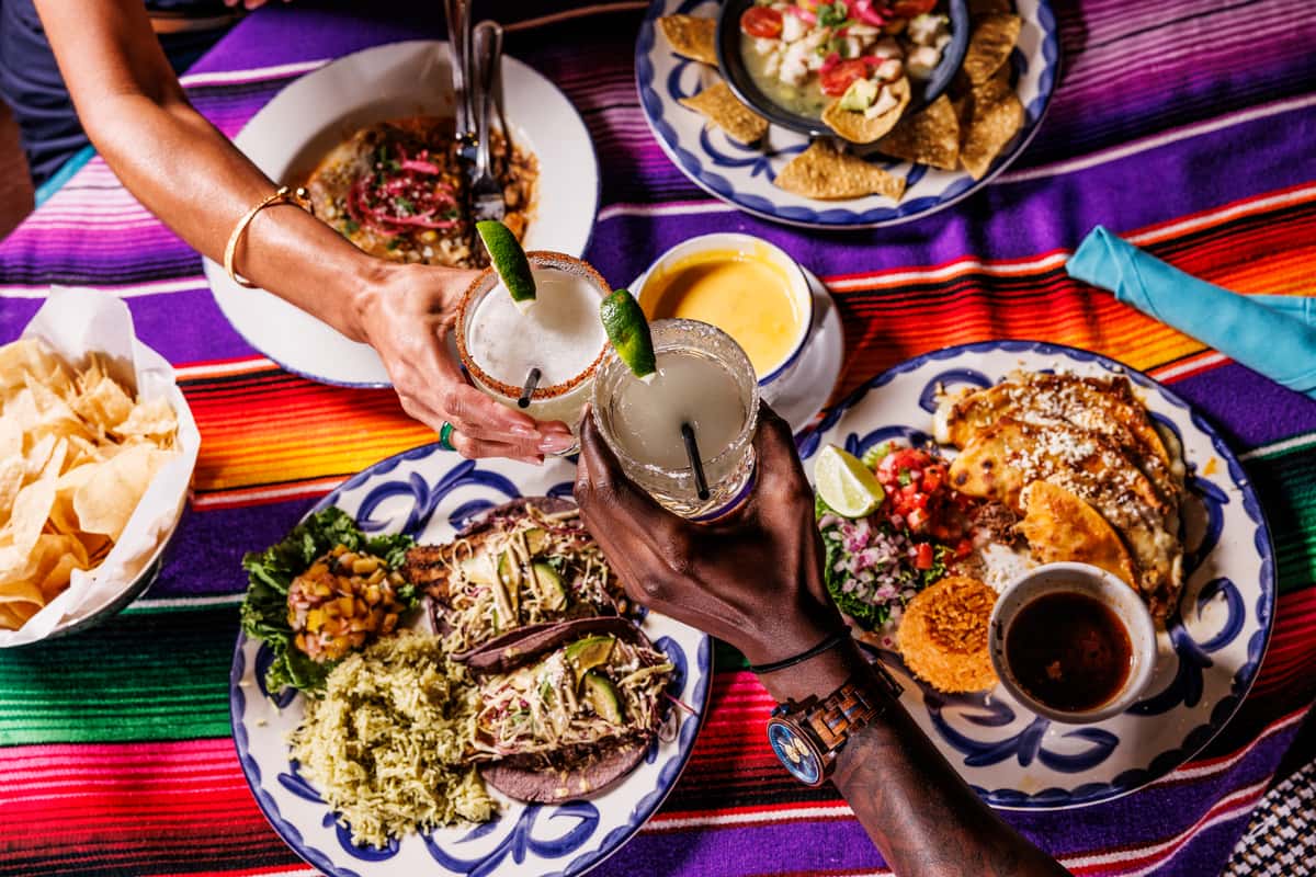 Vibrant Mexican feast with grilled fish tacos, birria tacos with rice and beans, ceviche, queso dip, and chips, as two people toast with lime-garnished margaritas over a colorful serape table setting.