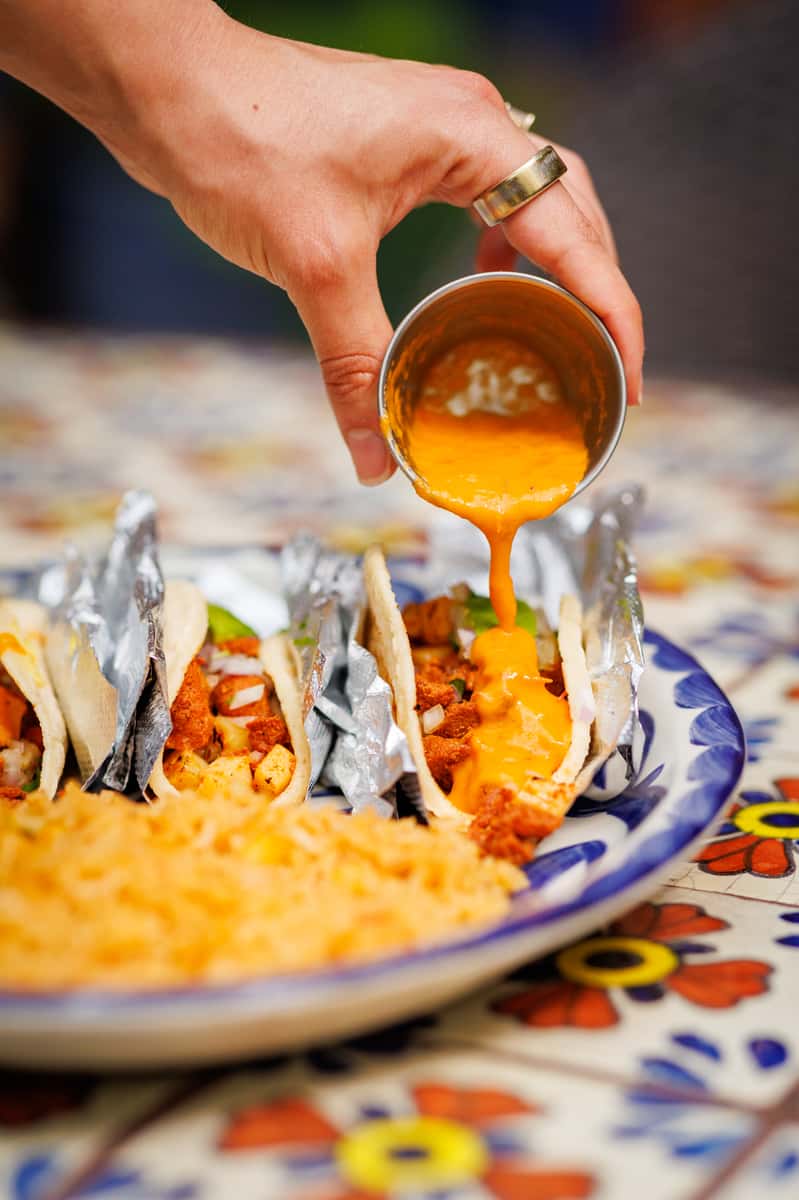 Hand pouring creamy queso sauce over soft tacos filled with seasoned meat, onions, and avocado, wrapped in foil and served with a generous portion of Mexican rice on a vibrant Talavera-patterned plate.