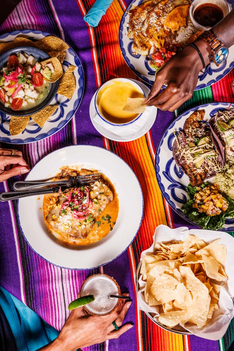 Vibrant Mexican feast on a colorful striped serape tablecloth, featuring creamy queso dip, fresh tortilla chips, ceviche with tomatoes and avocado, enchiladas, tacos with purple tortillas, and a creamy sauced entrée, accompanied by a salt-rimmed margarita with lime and friends enjoying each bite.