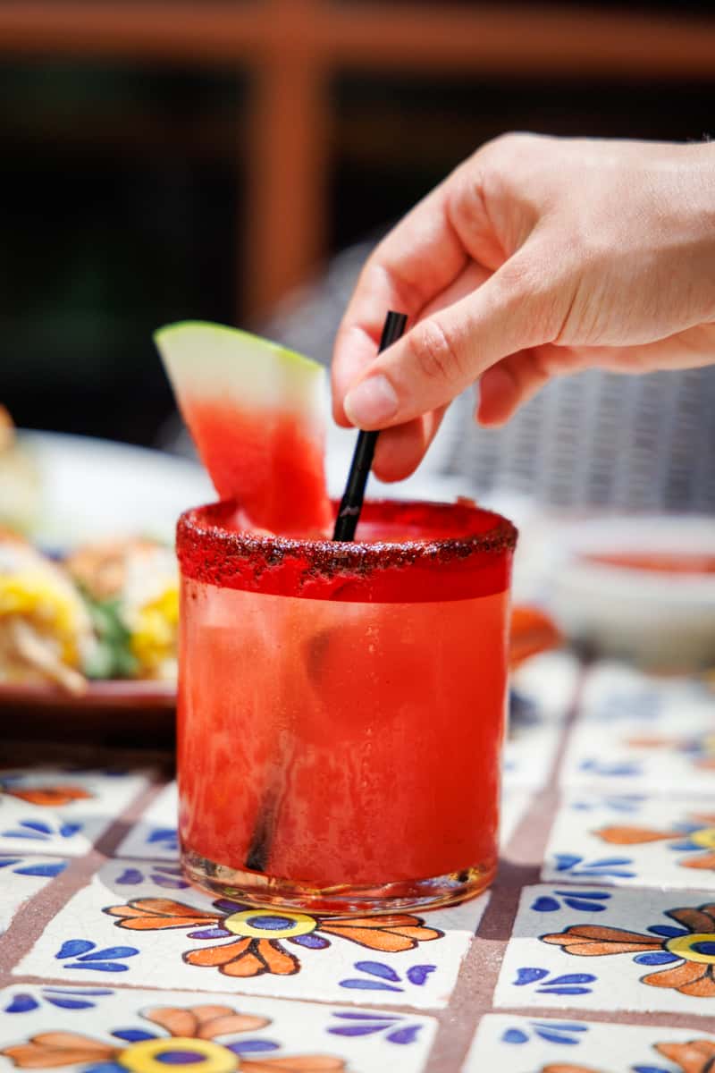 Refreshing watermelon margarita in a glass rimmed with chili salt, garnished with a fresh watermelon wedge, as a hand reaches to stir the drink—set on a festive Mexican tile table outdoors.