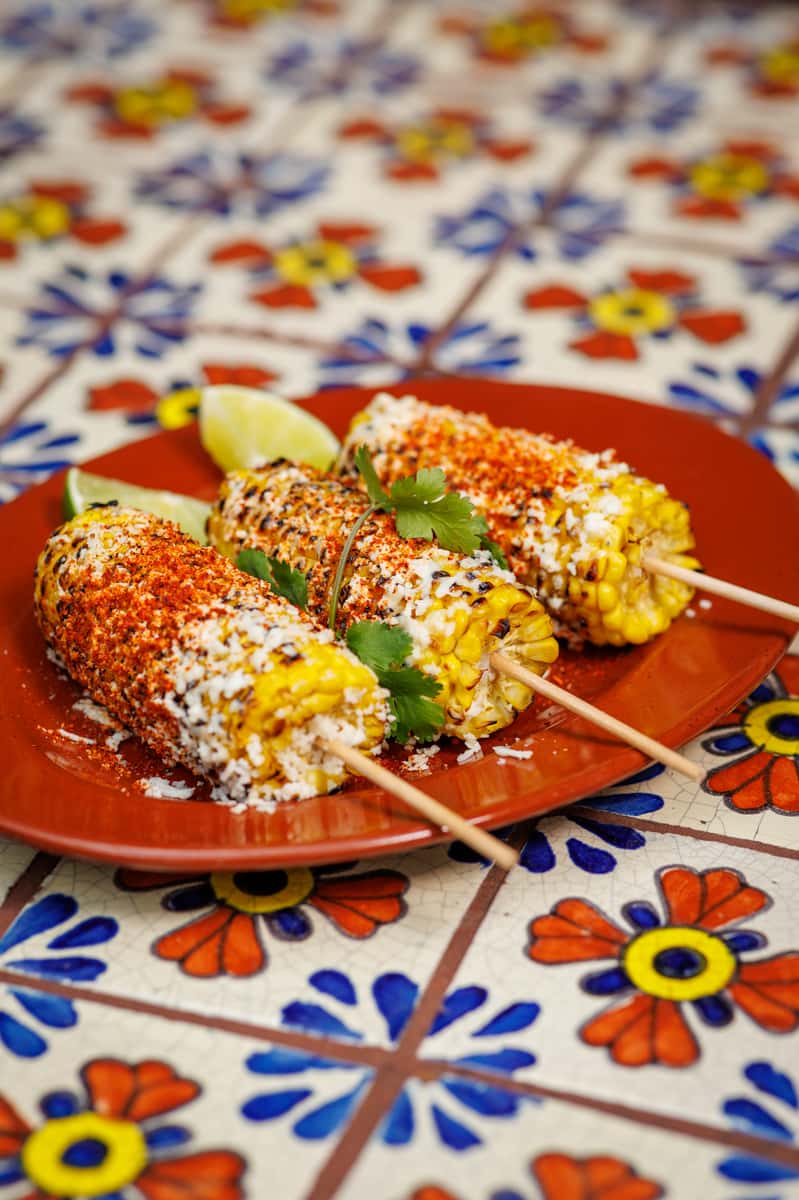 Authentic Mexican street corn (elotes) on wooden skewers, coated in cotija cheese, chili powder, and crema, served with lime wedges on a red plate atop a hand-painted Talavera tile table.