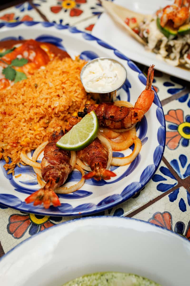 Grilled bacon-wrapped shrimp served on sautéed onions with a lime wedge, fluffy Mexican rice, and a side of crema on a blue-rimmed plate—perfect for a bold seafood entrée.