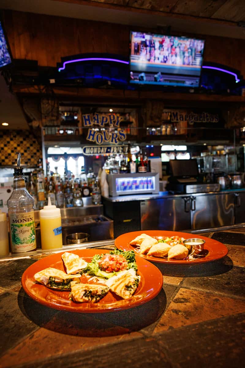 Warm bar setting featuring two plates of Mexican appetizers, including golden empanadas and grilled quesadillas with melted cheese and sautéed greens, served with fresh pico de gallo and dipping sauce. The background highlights a fully stocked bar, "Happy Hour" signage, and flat-screen TVs, creating a festive and casual dining atmosphere.