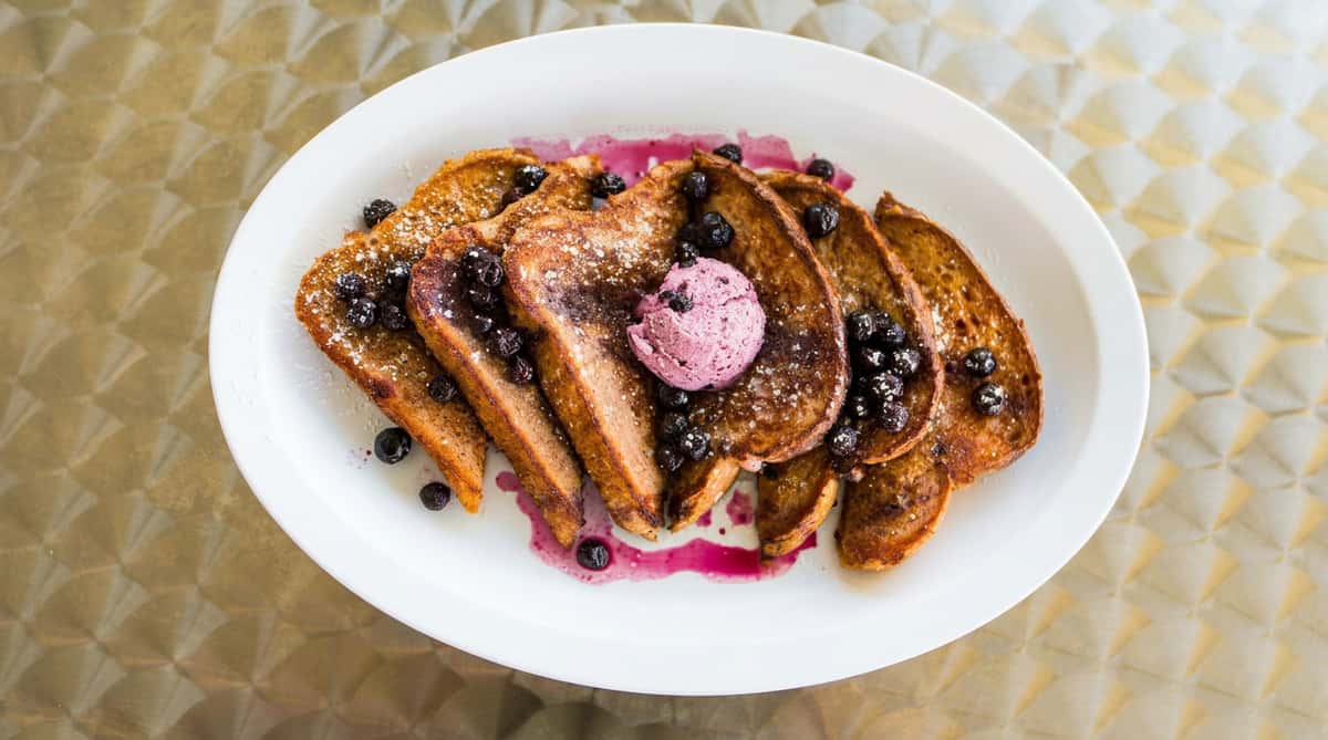 French toast slices with blueberries, powdered sugar, and a scoop of blueberry-swirl butter on a white plate.