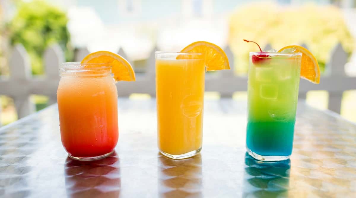 Three vibrant drinks on a table: orange-red gradient in a mason jar, orange juice, and a blue-green gradient with a cherry and orange slice.