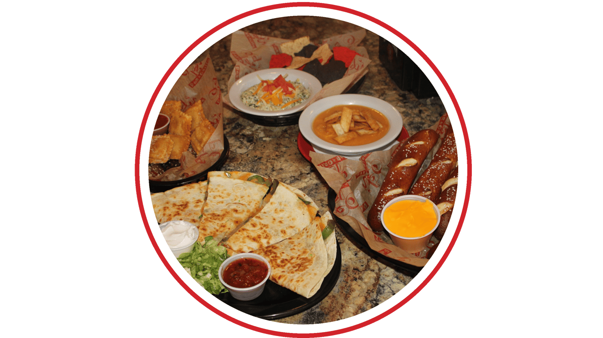 Assorted restaurant appetizers and snacks on a granite tabletop, including quesadillas, soft pretzels with cheese dip, tortilla chips with spinach dip, creamy soup, and fried pastries.