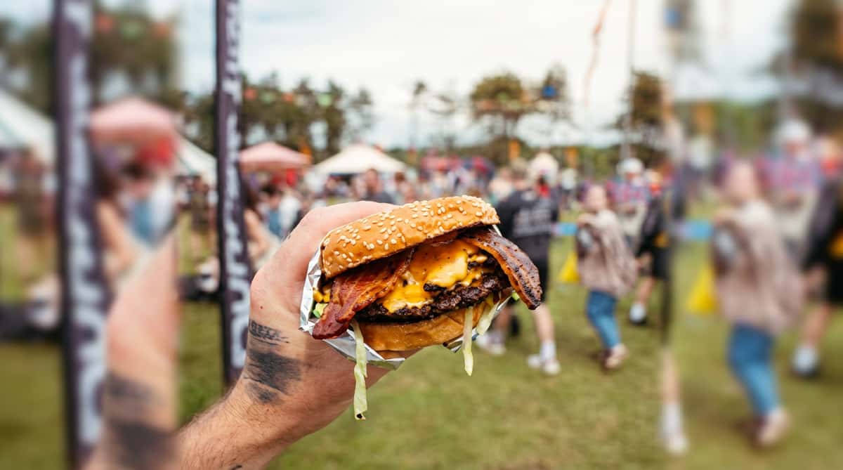 Hand holding a double cheeseburger with bacon and lettuce, wrapped in foil, against a blurry outdoor festival background.