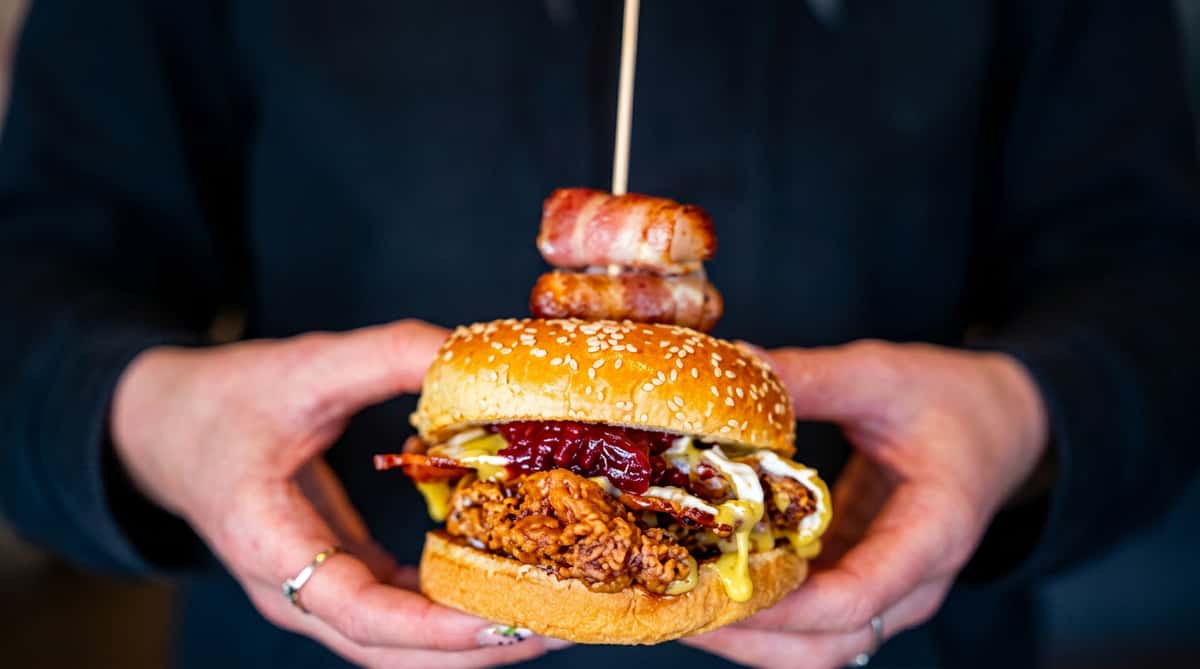 A large fried chicken sandwich with bacon, red jam, and sauce on a sesame bun, held by two hands.