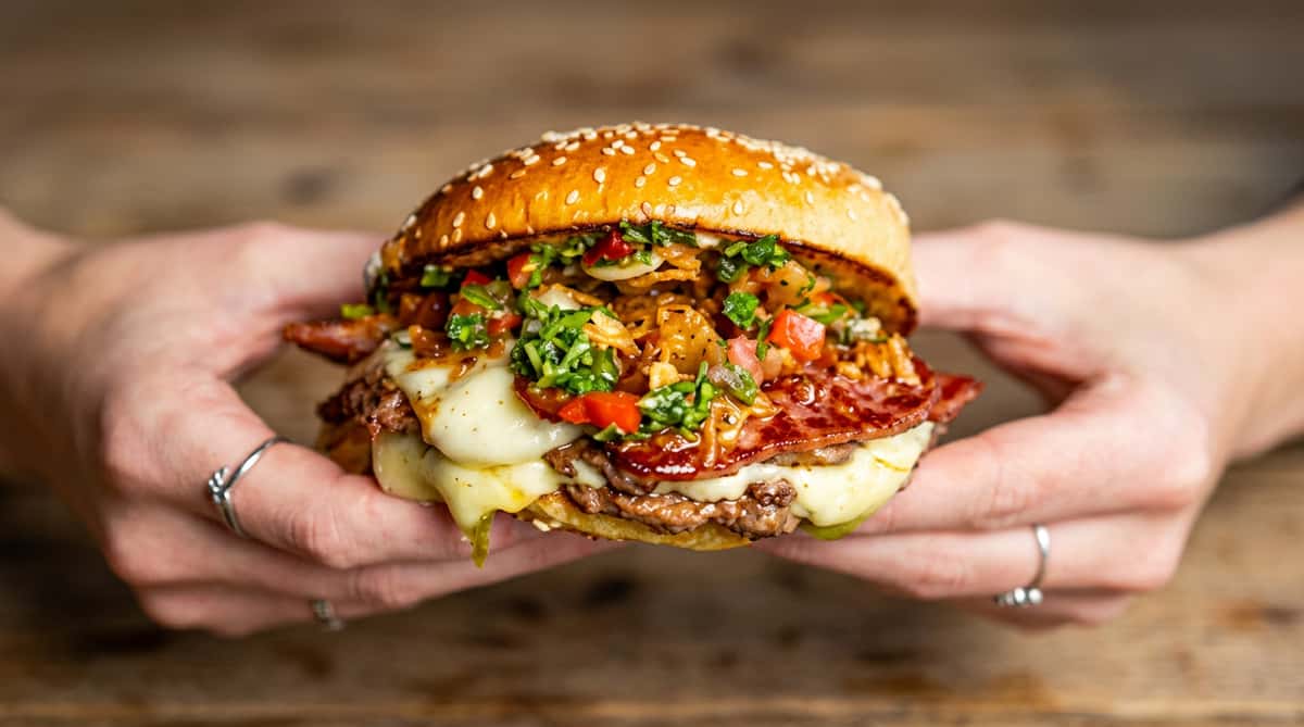 Two hands holding a large burger with a sesame seed bun, melted cheese, meat, and green relish.