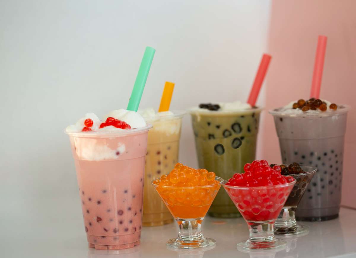 A close-up shot features an assortment of vibrant, colorful boba teas in clear plastic cups with straws. Three small glass bowls showcase different flavors of boba pearls, adding to the variety and visual appeal of the drink options.