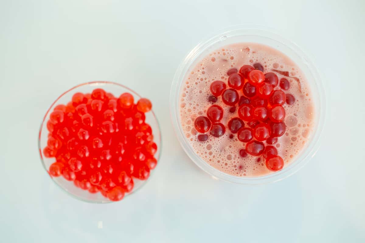 Red popping boba in a glass bowl, and on top of a pink drink. These are likely ingredients for bubble tea, a signature offering.