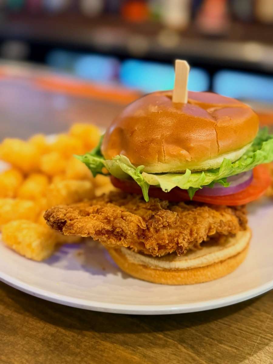 Crispy fried chicken sandwich topped with lettuce, tomato, and red onion on a toasted brioche bun, served with golden tater tots.