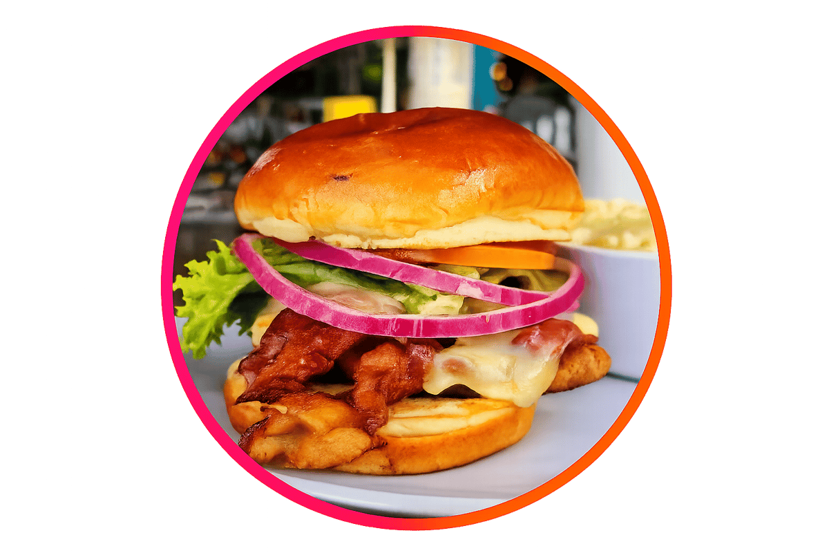 Close-up bacon cheeseburger with crispy bacon, melted cheese, fresh lettuce, tomato, red onion, and pickles on a toasted brioche bun, featured as a signature menu item at The Boxer Kitchen & Bar.
