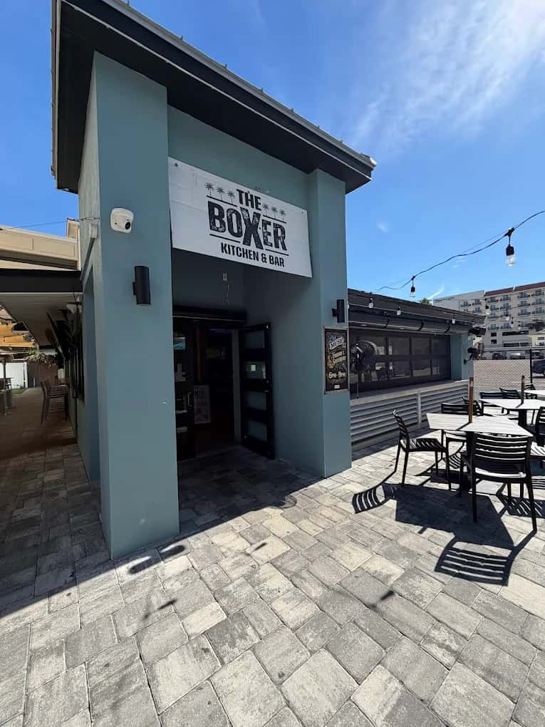 Exterior entrance of The Boxer Kitchen & Bar featuring modern coastal architecture, outdoor patio seating, and a casual waterfront dining atmosphere under a clear blue sky.
