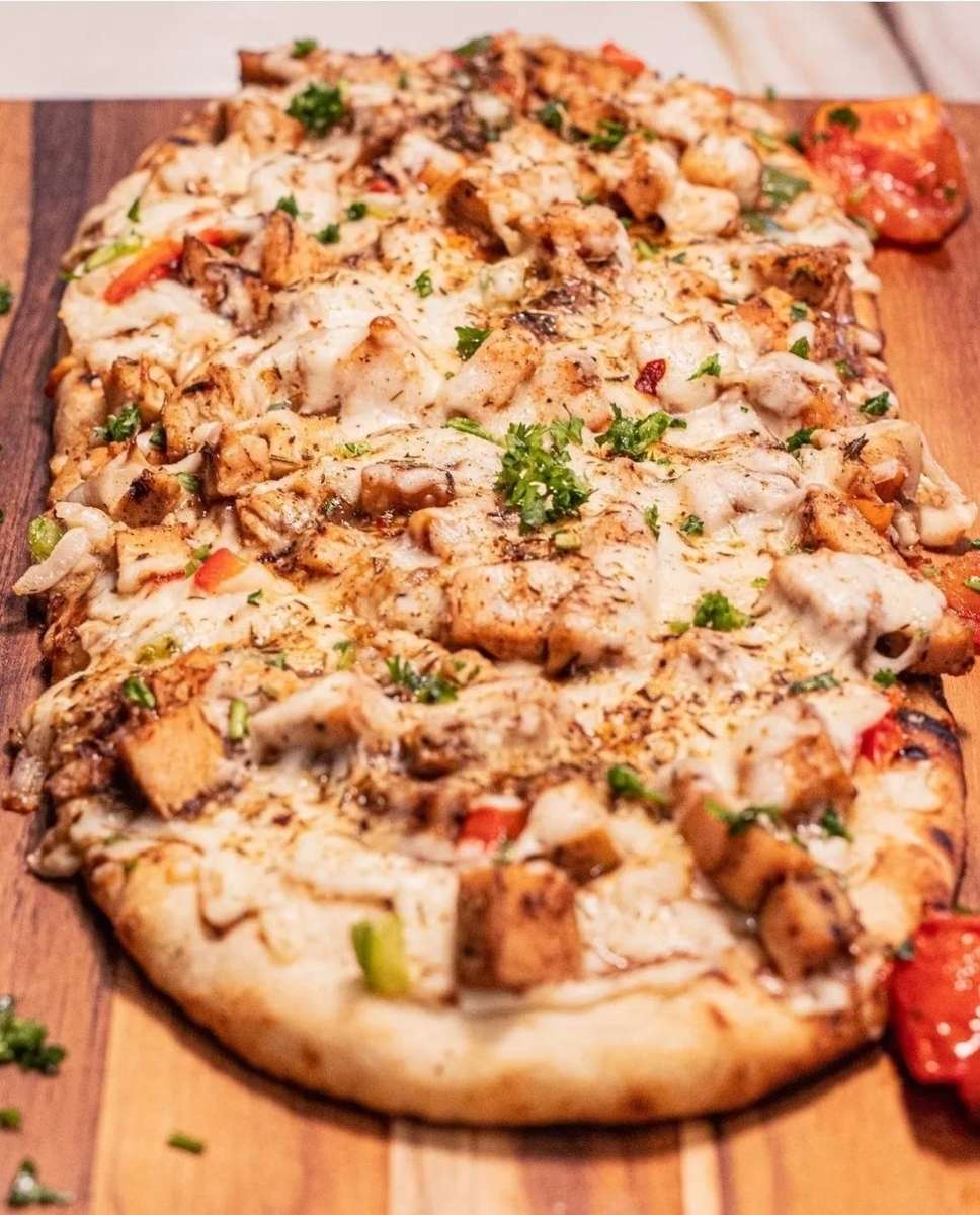 Flatbread pizza topped with grilled chicken, melted mozzarella, roasted red peppers, herbs, and seasoning on a wooden board