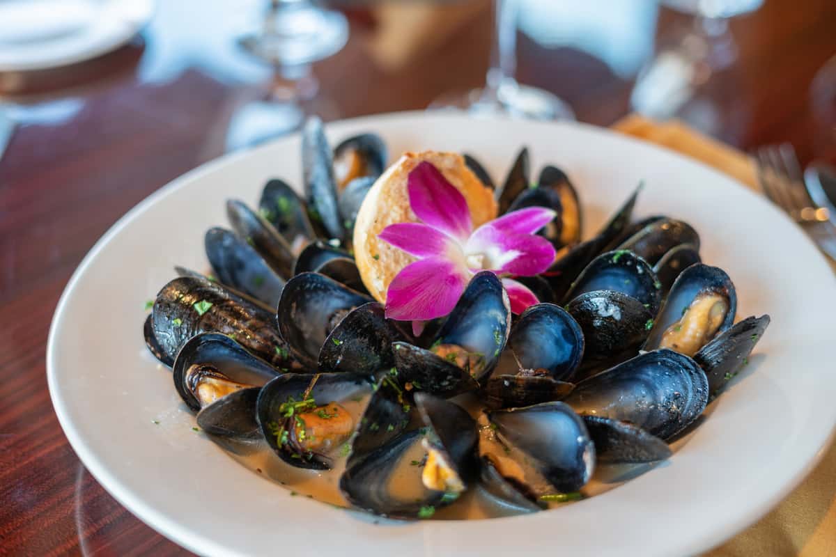 A flavorful seafood dish featuring fresh steamed mussels served in their shells, arranged in a generous bowl and coated in a rich, savory sauce that highlights their natural ocean flavor. The mussels are beautifully presented with a vibrant purple orchid garnish, adding a touch of elegance and color. This dish captures the essence of classic Italian coastal cuisine, combining simplicity, freshness, and bold Mediterranean flavors for a refined dining experience.