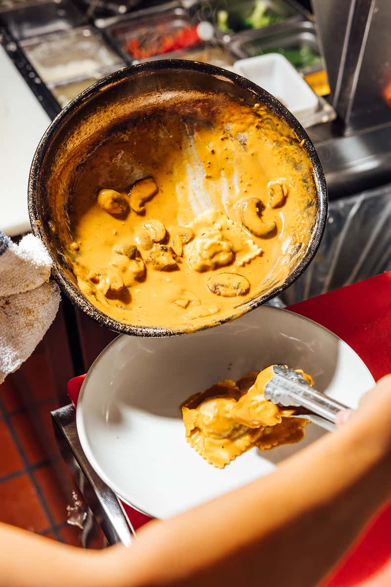Chef plating ravioli covered in a rich, creamy mushroom sauce straight from a sauté pan in a professional kitchen—highlighting the indulgent flavors and handcrafted preparation of classic Italian pasta dishes.