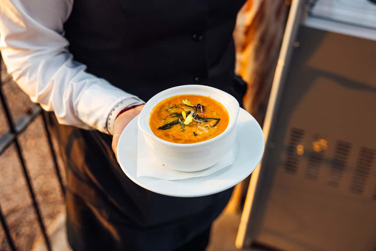 Server presenting a bowl of creamy tomato basil soup garnished with fresh herbs and microgreens, served in a white ceramic bowl on a patio—ideal for highlighting warm, comforting starters in an upscale Italian dining experience.