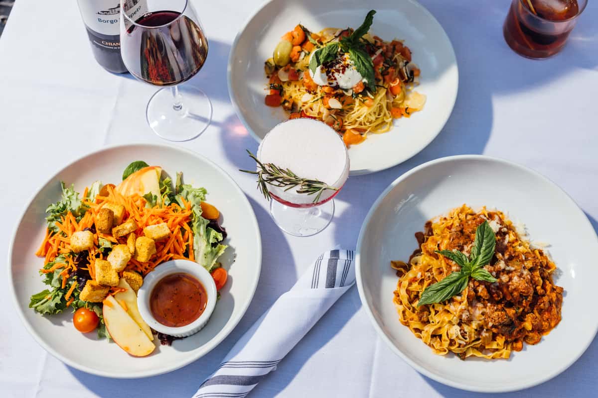 Outdoor Italian dining spread featuring a fresh garden salad with apple slices and croutons, creamy burrata pasta with cherry tomatoes and basil, rich tagliatelle Bolognese topped with parmesan and basil, and a rosemary-garnished cocktail, paired with red wine—ideal for showcasing authentic Italian cuisine and alfresco dining ambiance.