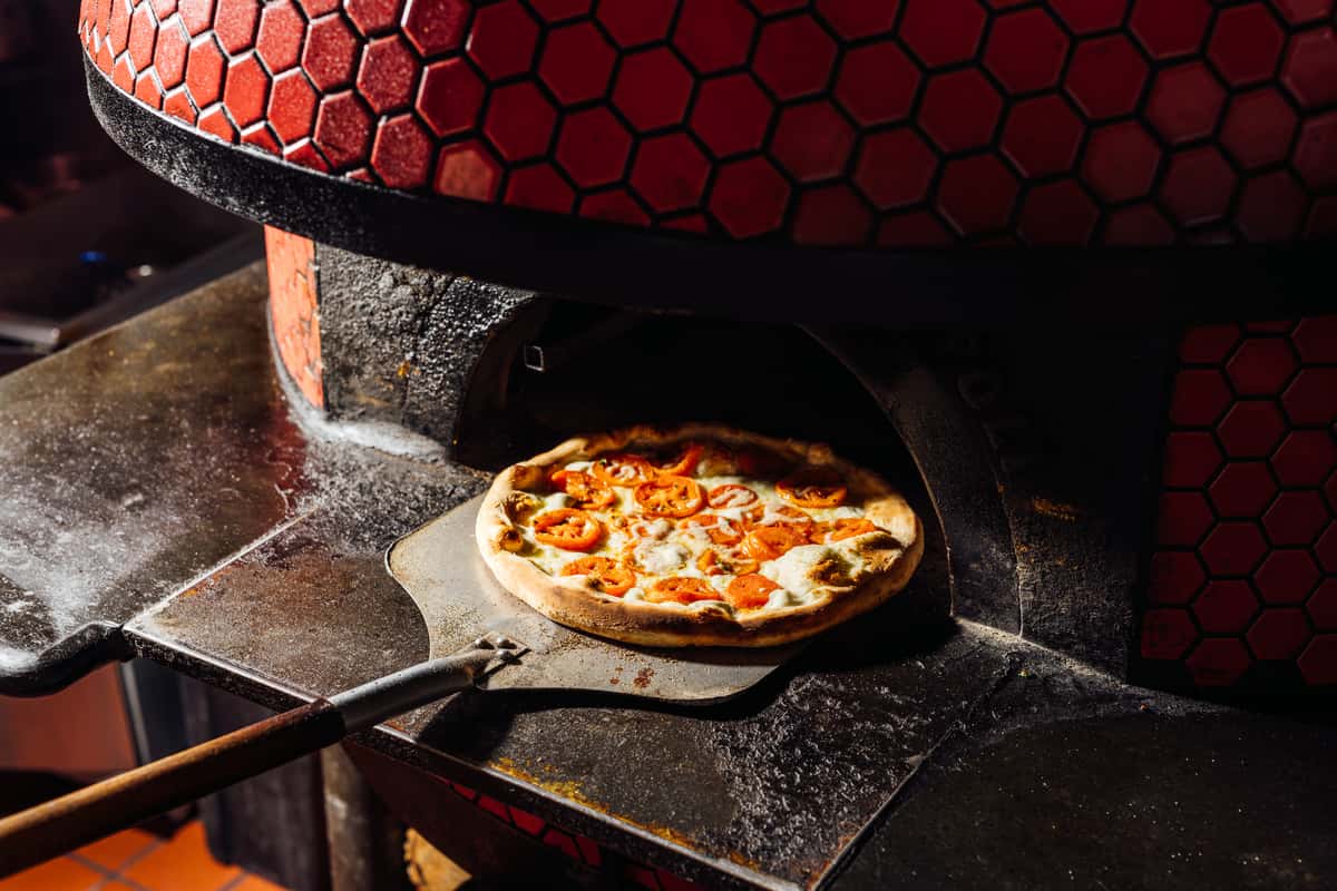 Freshly baked Margherita pizza with melted mozzarella and sliced tomatoes being pulled from a red-tiled wood-fired oven—perfect for showcasing authentic Neapolitan-style pizza and traditional Italian cooking techniques.