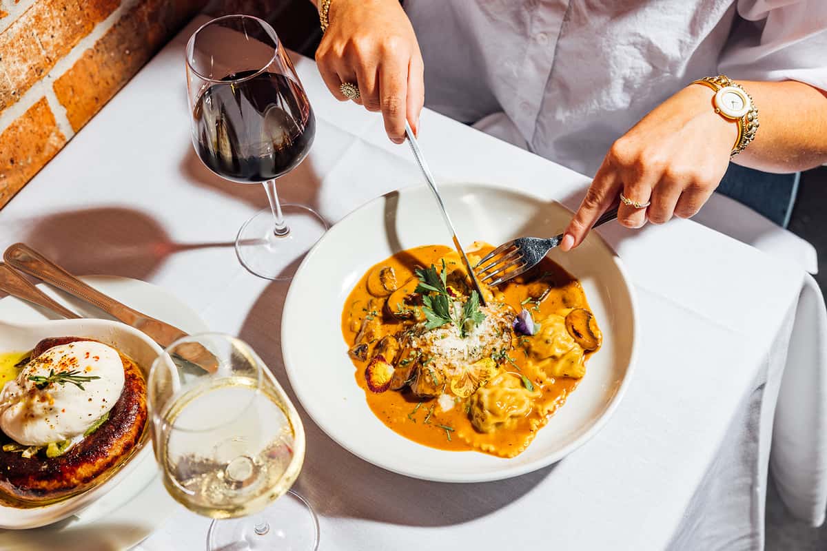 Elegant Italian pasta dish in creamy tomato sauce topped with shaved Parmesan and fresh herbs, served on a white plate alongside red and white wine glasses in a cozy brick-walled restaurant setting.