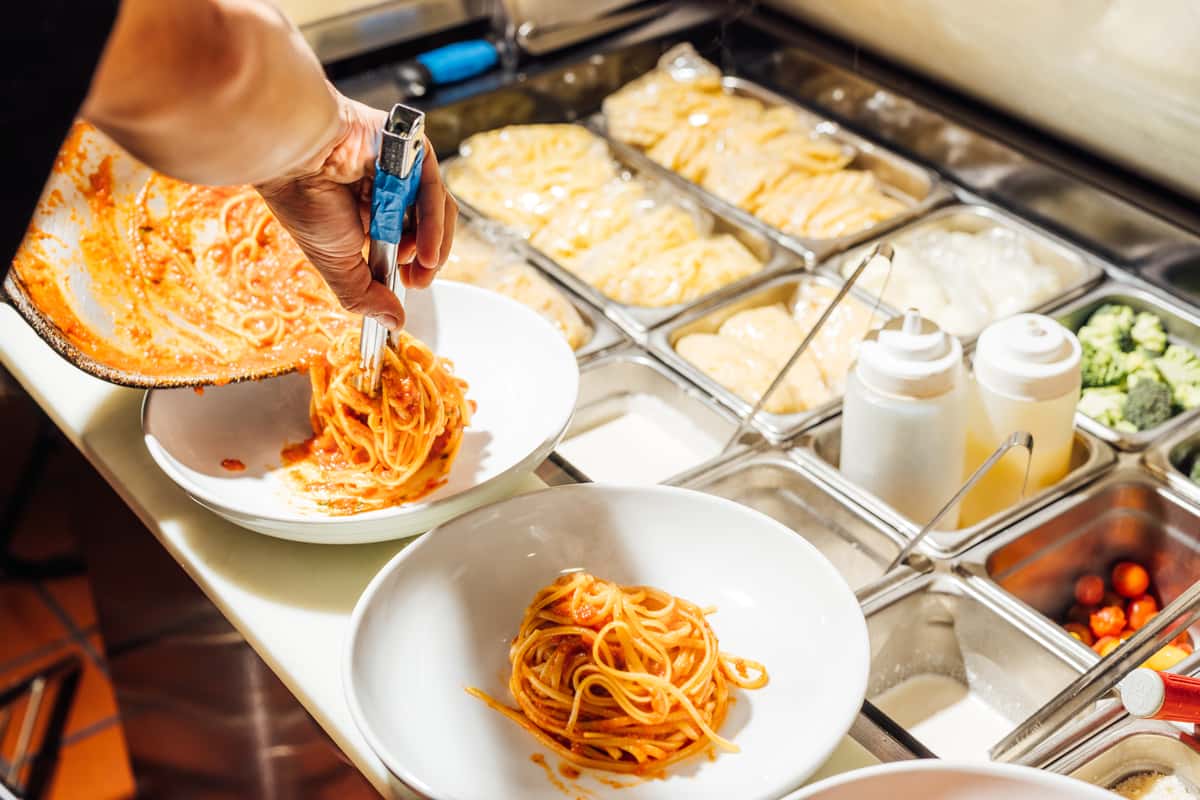 Chef plating freshly cooked spaghetti tossed in tomato sauce in a professional Italian kitchen, with various pasta types and ingredients visible in stainless steel prep containers—highlighting authentic pasta preparation and handcrafted Italian cuisine.