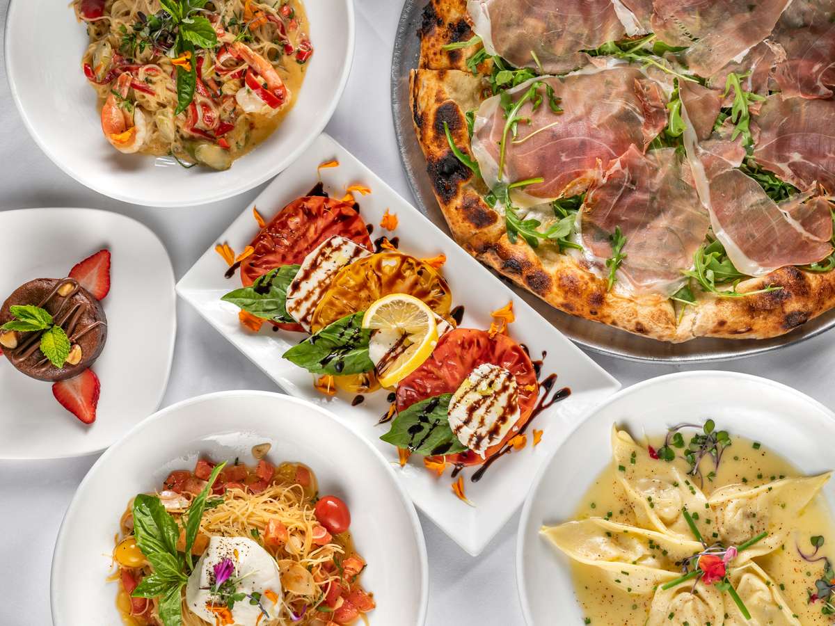 Top-down view of an elegant Italian spread featuring prosciutto and arugula pizza, heirloom tomato and mozzarella caprese salad, burrata pasta with cherry tomatoes, seafood pasta, cheese-filled ravioli in cream sauce, and a chocolate dessert with strawberries—perfect for showcasing gourmet Italian cuisine in a fine dining setting.